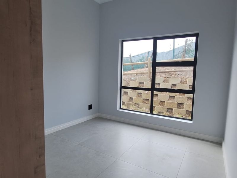 A clean, empty bedroom featuring modern grey floor tiles, neutral walls, and a large black aluminium window overlooking the rear exterior.