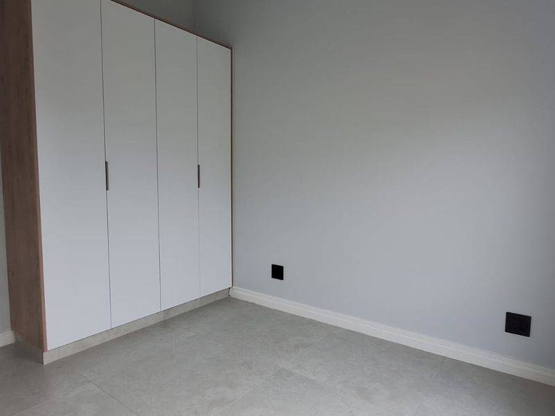 A bright, modern bedroom featuring neutral grey walls, tiled flooring, and sleek built-in cupboards with a wood-grain finish.