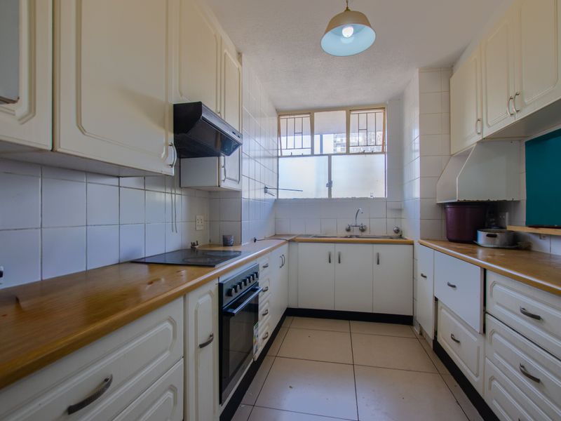 A functional galley-style kitchen featuring white cabinetry, laminate countertops, built-in oven, electric hob, and tiled finishes throughout.