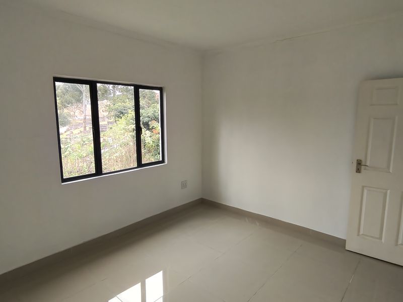 A bright, empty bedroom featuring neutral tiled flooring, white walls, and a black-framed window overlooking the exterior.