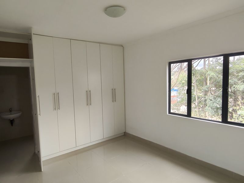 A bright, empty bedroom featuring built-in cupboards, tiled flooring, and large windows with a view of the surrounding trees.