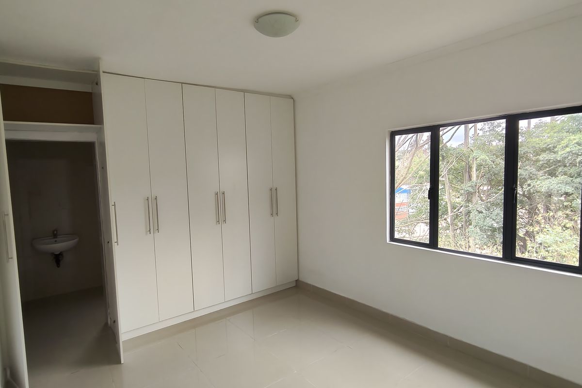 A bright, empty bedroom featuring built-in cupboards, tiled flooring, and large windows with a view of the surrounding trees.