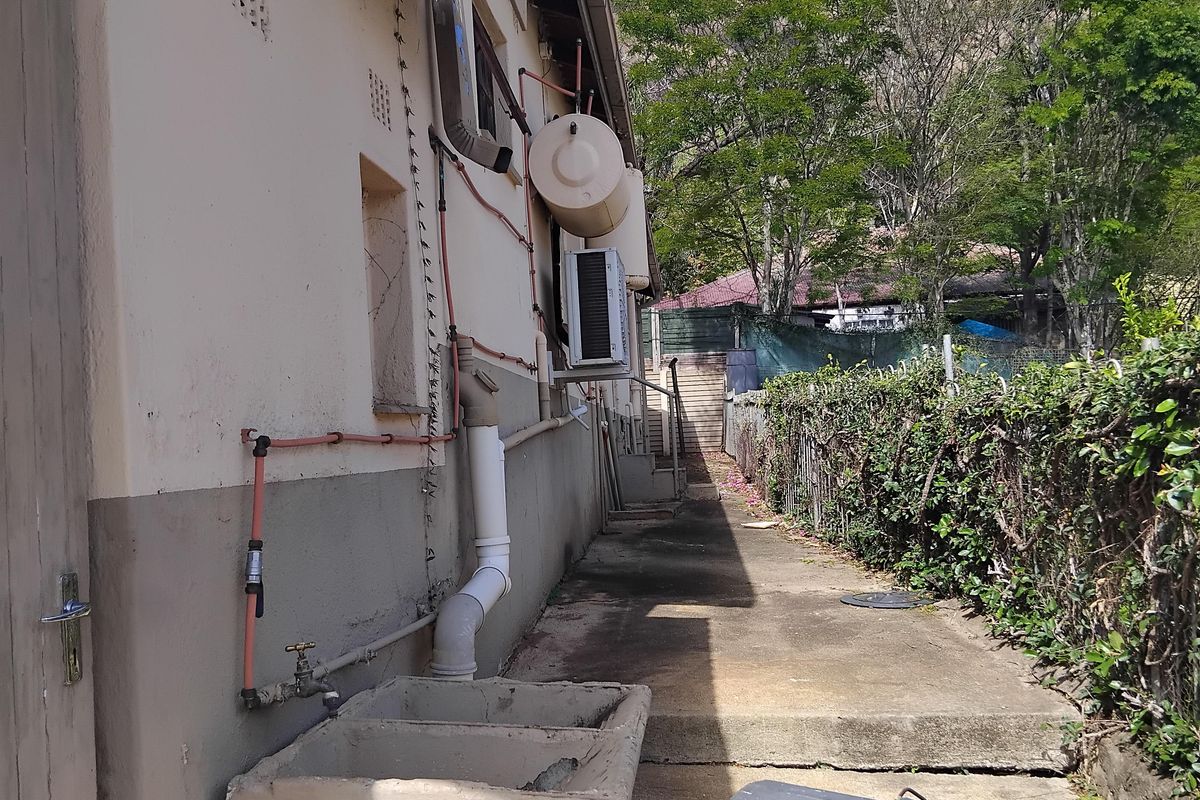 Rear exterior view showing a concrete pathway, external wash trough, geyser, and air conditioning unit along the side of the house.