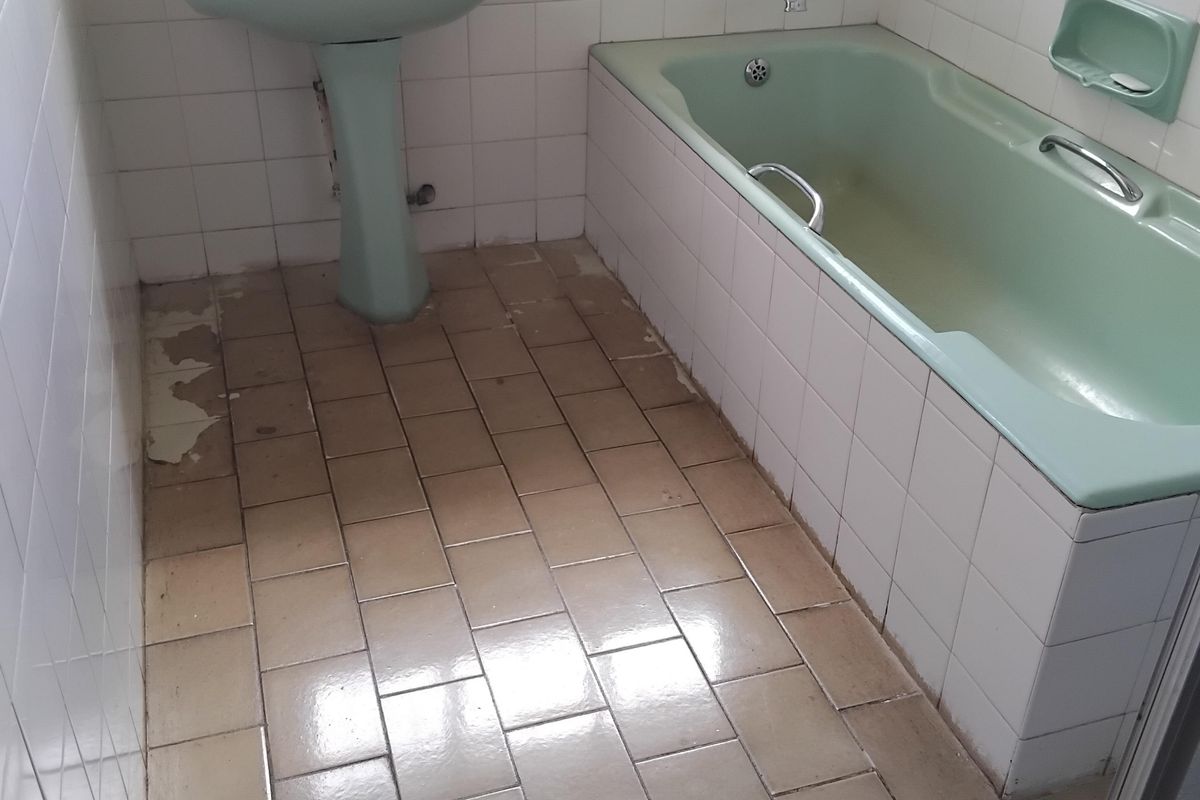A dated bathroom featuring a pastel green pedestal basin and matching bathtub, with white wall tiles and worn ceramic floor tiling.
