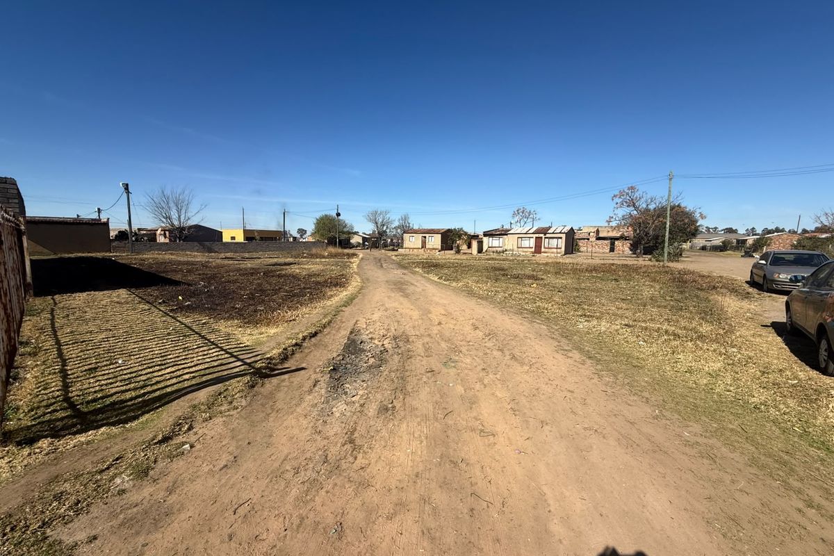 A view of an unpaved residential street with surrounding vacant land and utility infrastructure in a developing neighborhood.