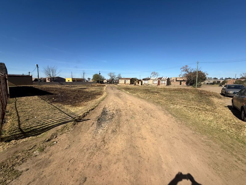 A view of an unpaved residential street with surrounding vacant land and utility infrastructure in a developing neighborhood.