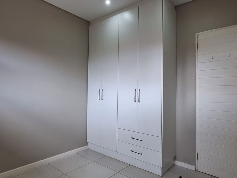 A clean, modern bedroom featuring neutral wall tones, tiled flooring, and a large white built-in wardrobe with integrated drawers.