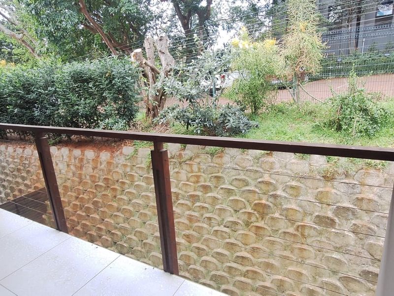 A private balcony featuring tiled flooring and a metal railing, overlooking a landscaped garden area with a stone retaining wall.