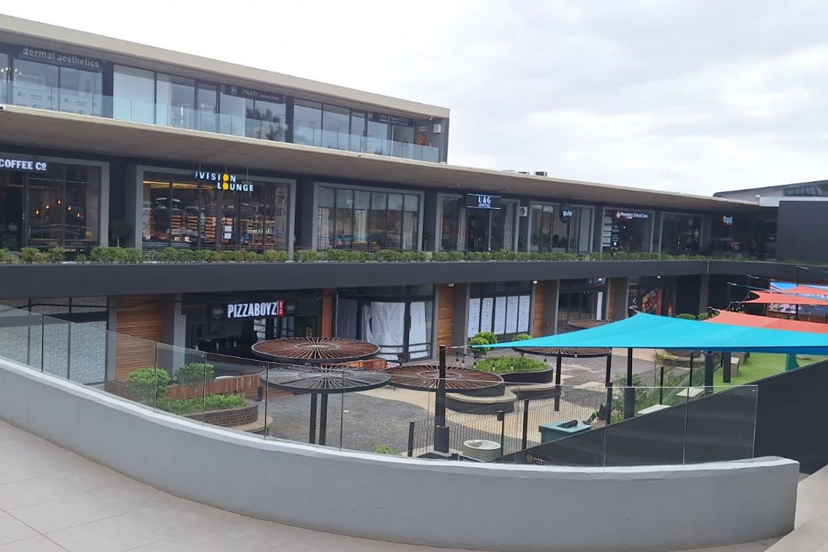 Modern multi-level commercial complex featuring glass-fronted retail units, a central paved courtyard with shade sails, and contemporary architectural finishes.