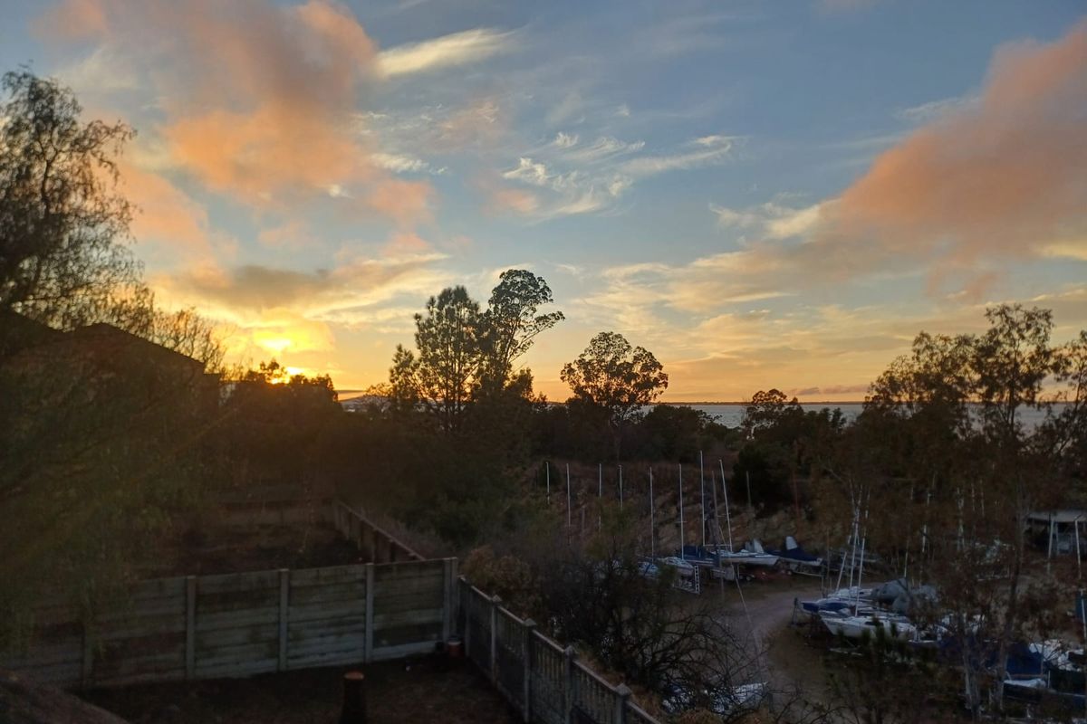 A scenic sunset view overlooking a coastal area with trees and a fenced yard in the foreground.