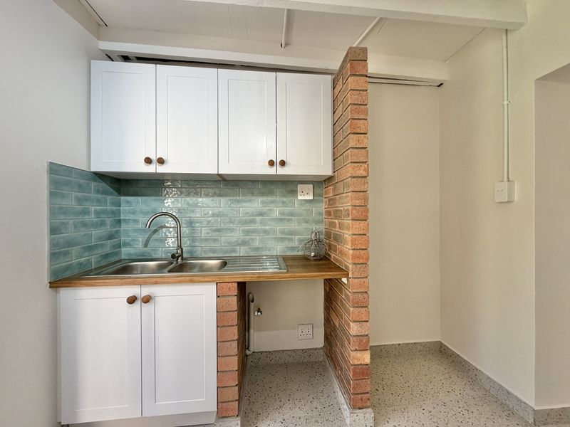 A compact, well-maintained kitchen area featuring white cabinetry, a blue tiled backsplash, timber countertops, and a distinctive exposed brick partition.
