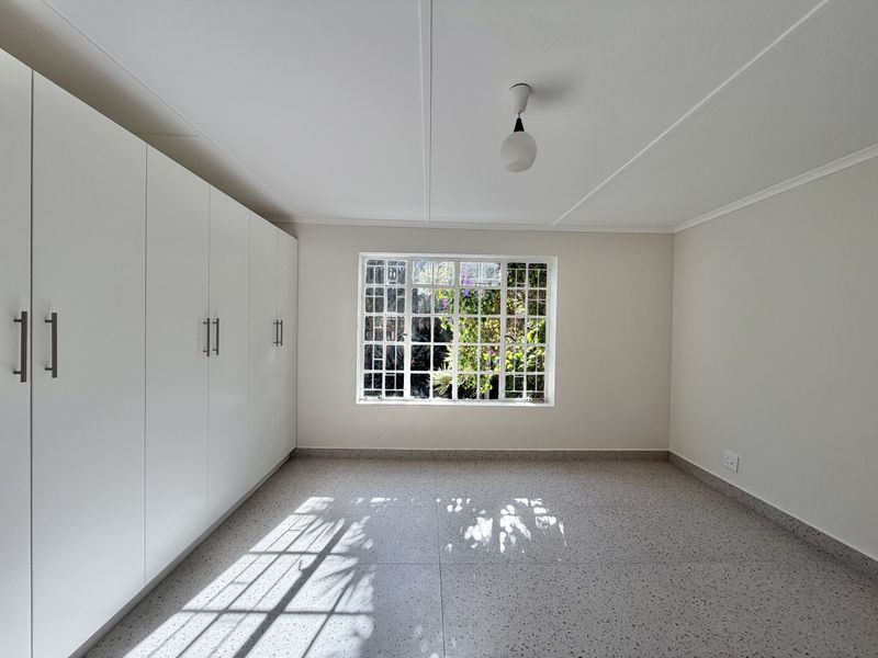 A bright, empty bedroom featuring built-in cupboards, terrazzo flooring, and a window with security bars overlooking the garden.