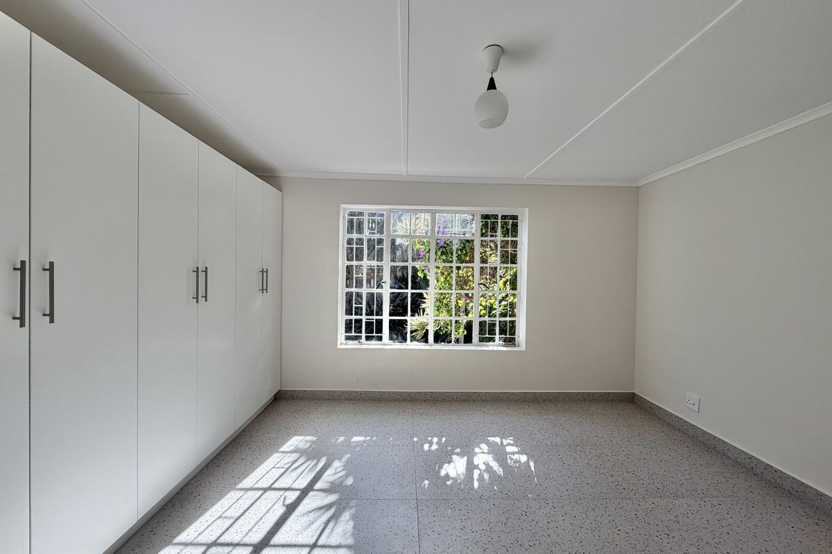 A bright, empty bedroom featuring built-in cupboards, terrazzo flooring, and a window with security bars overlooking the garden.