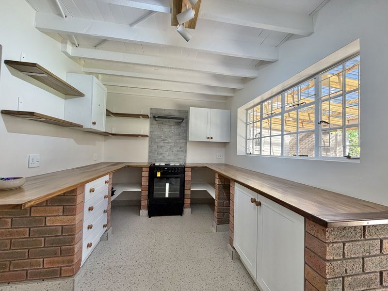 A bright, rustic-style kitchen featuring exposed white beams, brick-based cabinetry with wooden countertops, and a freestanding oven with a tiled splashback.