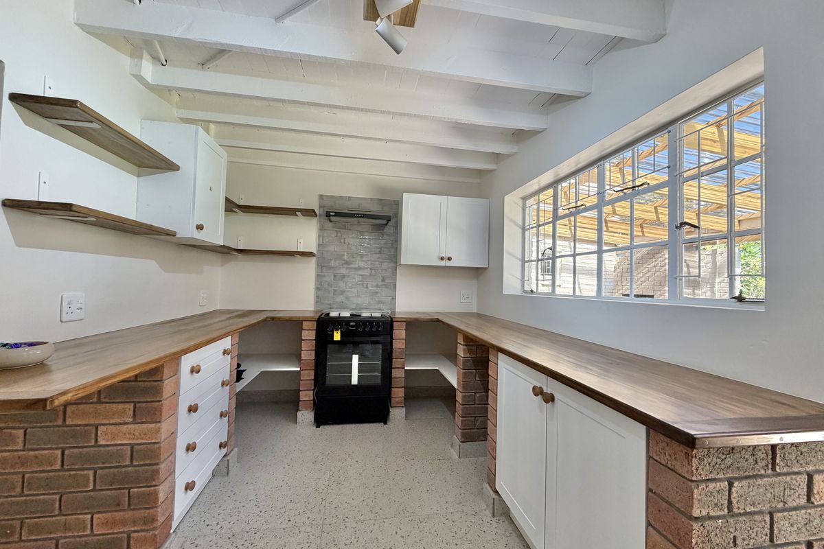 A bright, rustic-style kitchen featuring exposed white beams, brick-based cabinetry with wooden countertops, and a freestanding oven with a tiled splashback.
