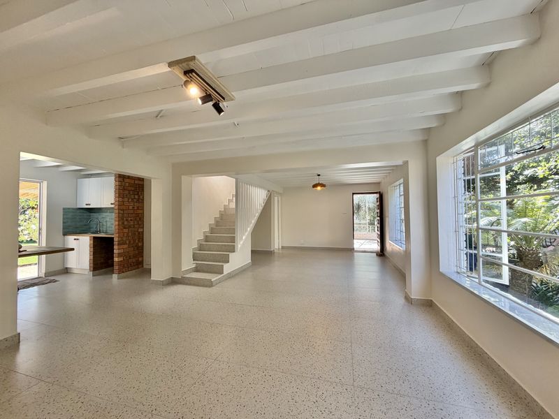 Spacious open-plan living area featuring exposed timber ceiling beams, terrazzo flooring, an exposed brick feature, and an integrated kitchen with direct garden access.