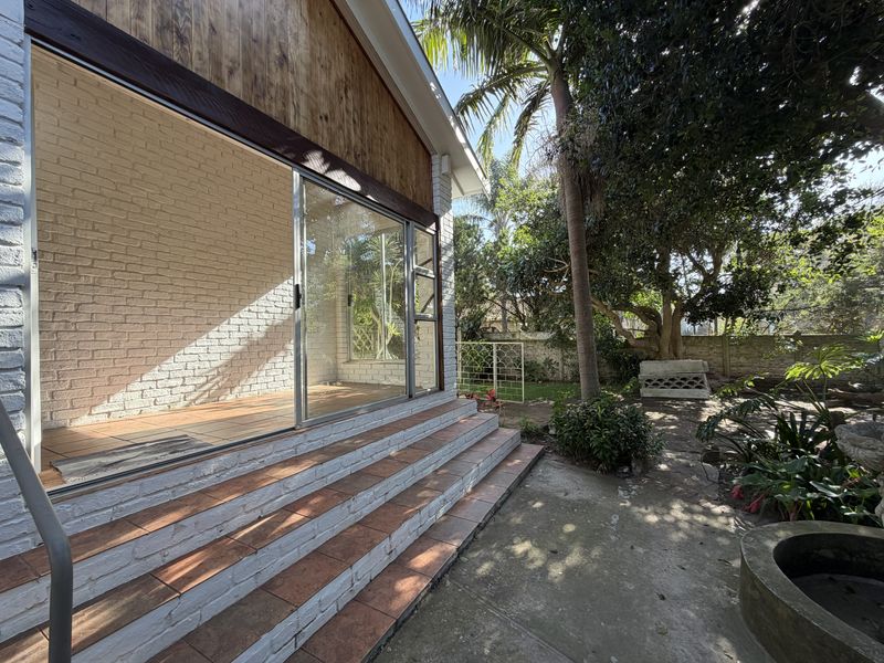 Rear exterior view featuring a covered patio with sliding glass doors, tiled steps, and access to a landscaped garden area.