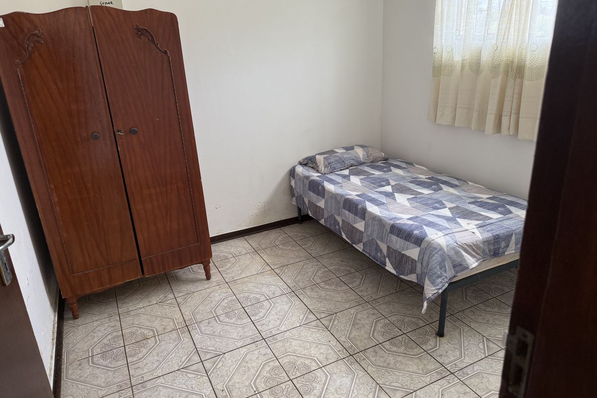 A simple bedroom featuring tiled flooring and a window with a curtain rail, offering a functional space ready for cosmetic updates.