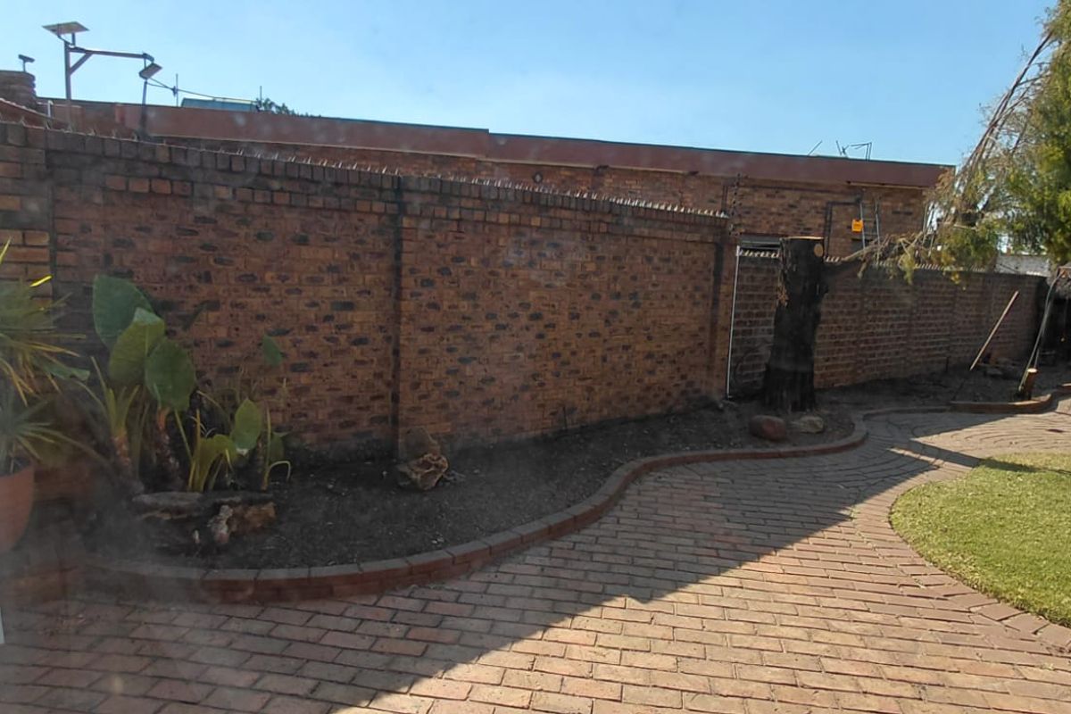 A rear garden area featuring a brick boundary wall with security spikes, paved pathways, and established garden beds.