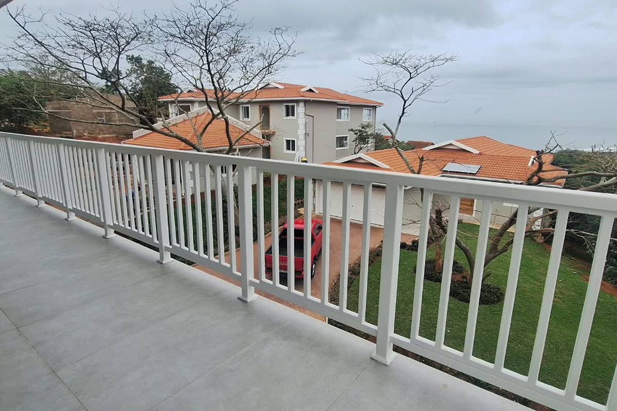 A spacious balcony featuring tiled flooring and a white metal balustrade, offering elevated views of the surrounding estate and coastal landscape.