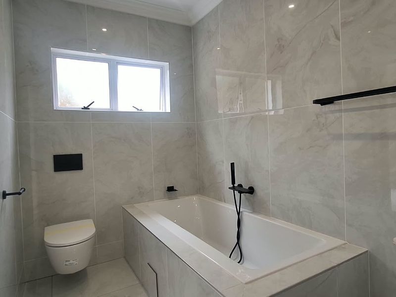 Modern bathroom featuring a built-in bathtub, wall-mounted toilet, stone-topped vanity, and contemporary black matte fixtures with neutral marble-look wall tiling.
