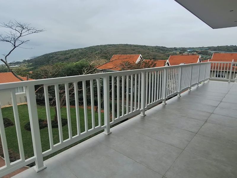 Spacious covered balcony featuring modern grey floor tiles and a white metal balustrade, offering elevated views of the surrounding estate and coastline.