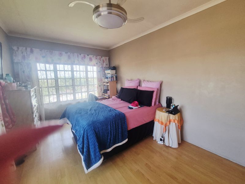 A bedroom featuring laminate flooring, a ceiling fan, and a large window with security bars, offering a functional space for rest.