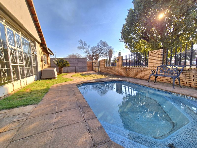 Outdoor pool area featuring a paved surround, brick boundary wall with metal fencing, and a well-maintained lawn.