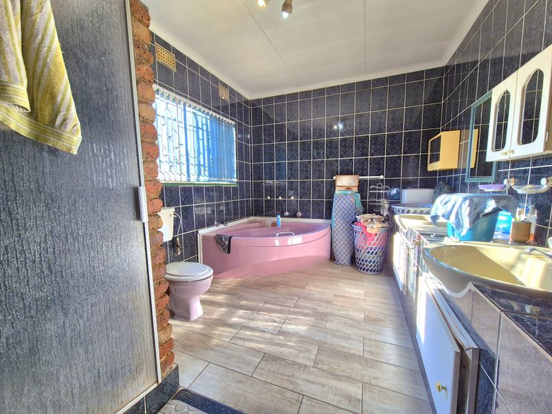 A dated bathroom featuring dark wall tiles, a pink corner bathtub, and a built-in vanity unit with a basin.