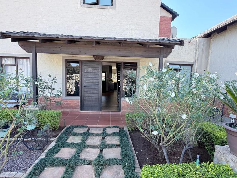 Rear exterior view featuring a covered patio with a timber pergola, stone paver walkway, and established garden beds leading to the main entrance.