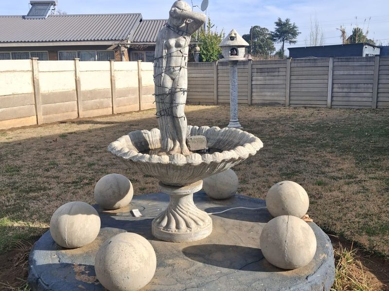 A backyard garden featuring a stone fountain with a statue, surrounded by concrete spheres and enclosed by a precast concrete boundary wall.