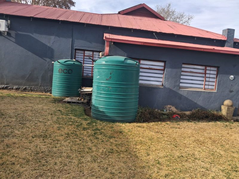 Rear exterior view of the house featuring a corrugated metal roof, brick chimney, and two large water storage tanks installed against the wall.