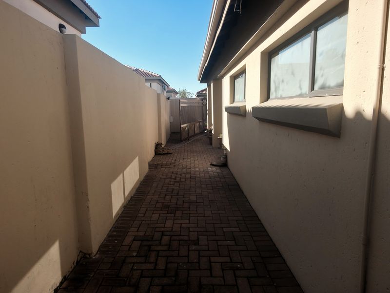 A narrow paved side passage between the house exterior and the boundary wall, featuring clean masonry and aluminium window frames.