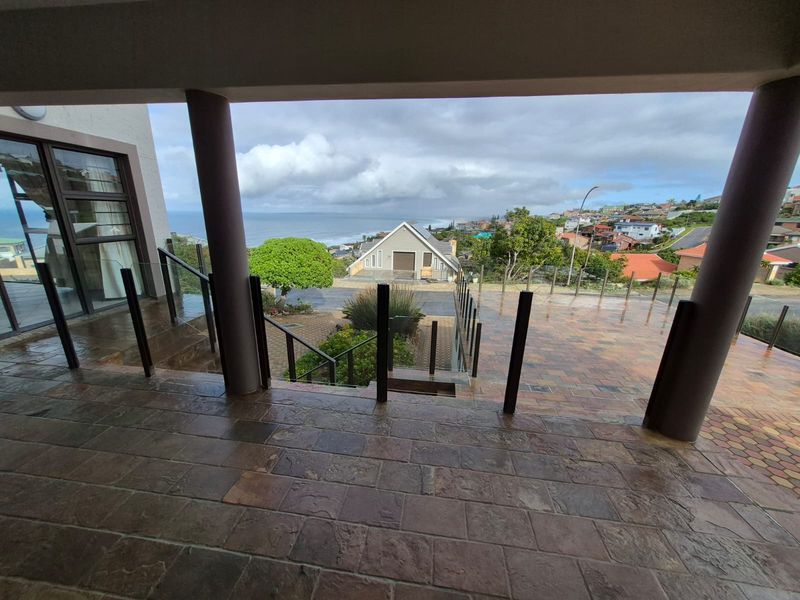 A spacious covered patio featuring stone tiling, glass balustrades, and structural columns, offering an elevated view of the surrounding coastal neighborhood.
