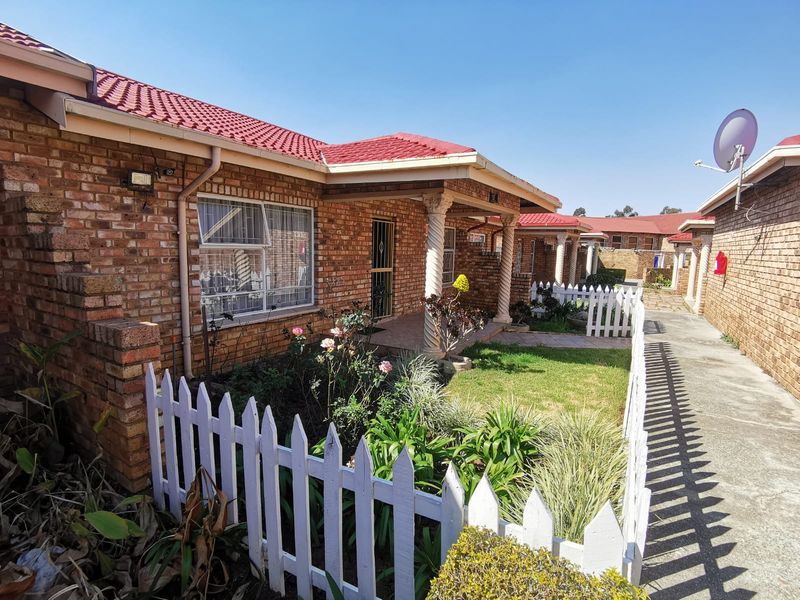 A well-maintained townhouse exterior featuring a face brick facade, tiled roof, covered porch with decorative columns, and a charming front garden with picket fencing.