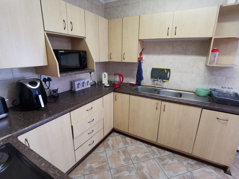 A functional kitchen featuring light wood-tone cabinetry, dark laminate countertops, a double stainless steel sink, and tiled backsplash with ceramic flooring.