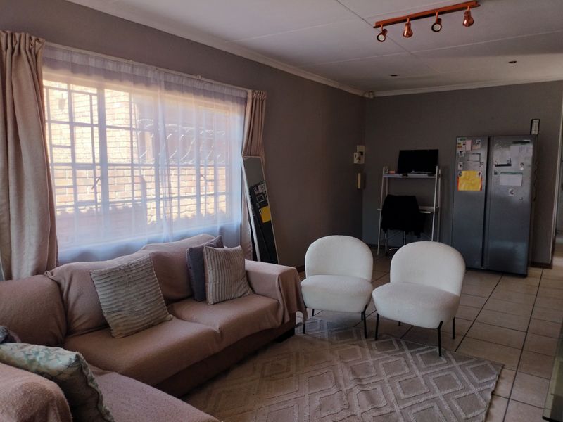 A living room area featuring tiled flooring, track lighting, and a large window with security bars, offering a functional and neutral living space.