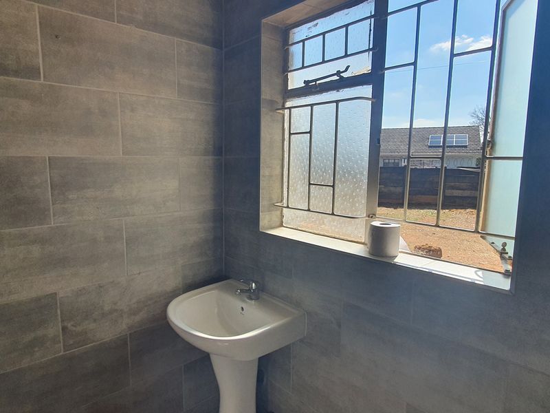 A compact bathroom featuring modern grey ceramic wall tiles, a pedestal basin with a mixer tap, and a window fitted with security bars.