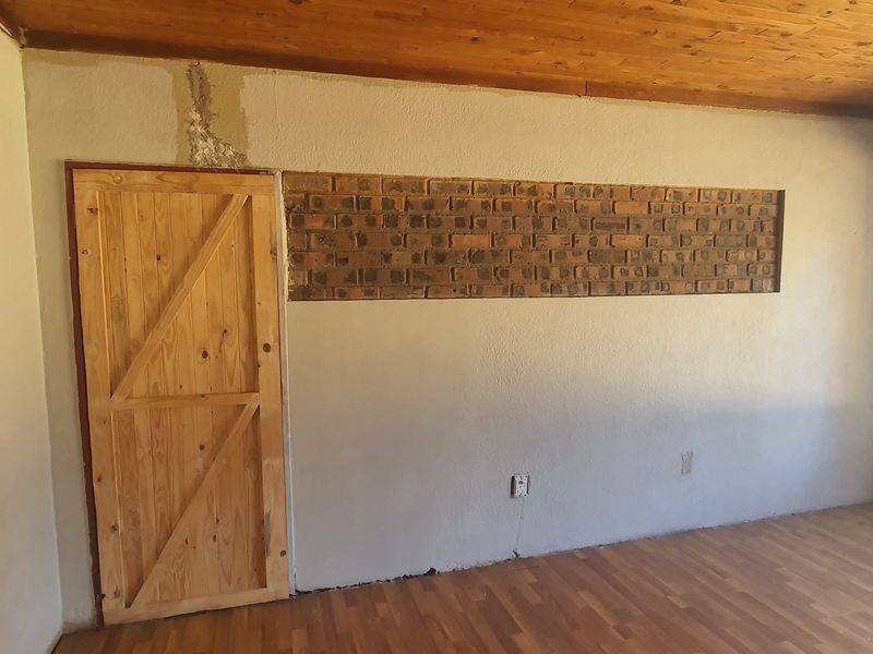 An empty living room featuring a timber ceiling, laminate flooring, and an exposed brick accent wall, requiring minor cosmetic finishing to the plasterwork.
