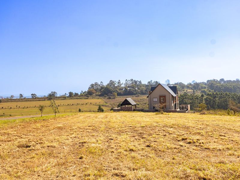 A wide-angle view of a vacant residential plot featuring a modern house in the distance, surrounded by open grassy fields and natural landscape.