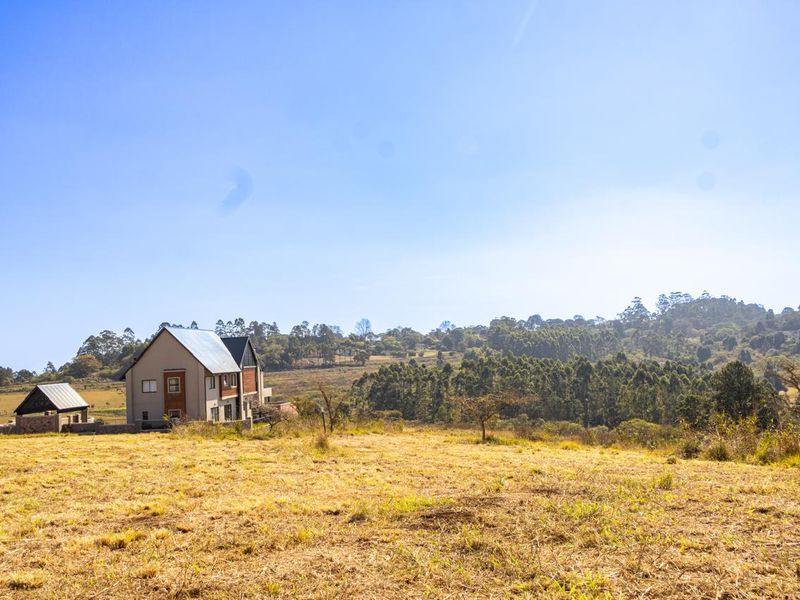 A wide-angle view of a vacant residential plot featuring natural grass terrain, surrounding trees, and a distant view of a modern residential property.