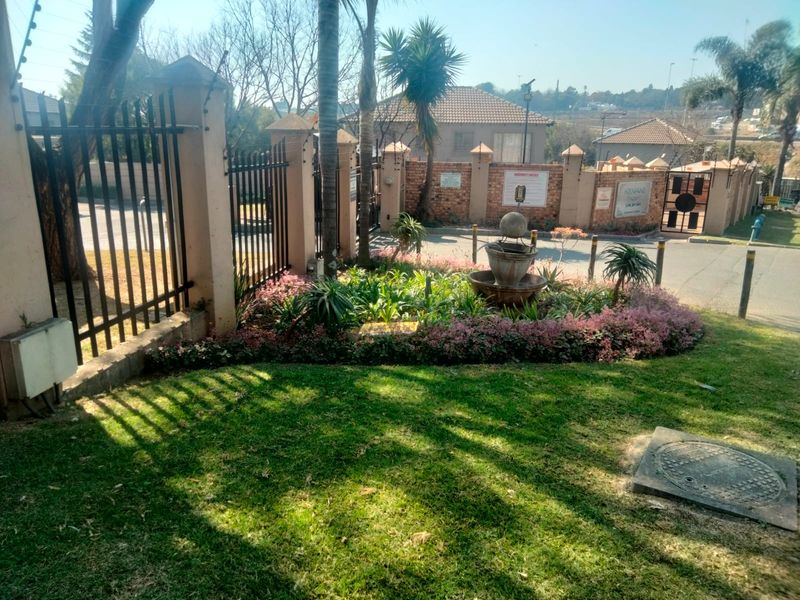 Front entrance area of the townhouse complex featuring landscaped garden beds, a decorative stone fountain, and secure perimeter fencing with electric security wires.