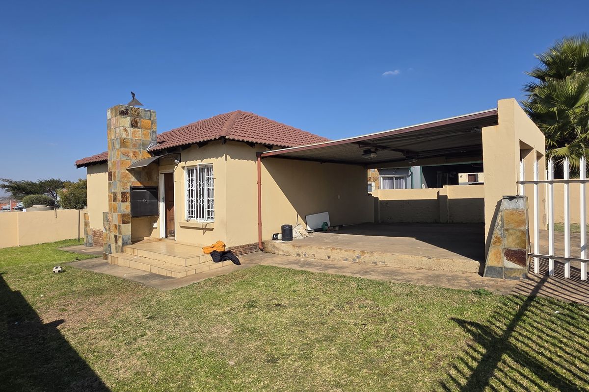 Rear view of the property featuring a covered patio, stone-clad chimney, and a well-maintained garden enclosed by a perimeter wall with security features.
