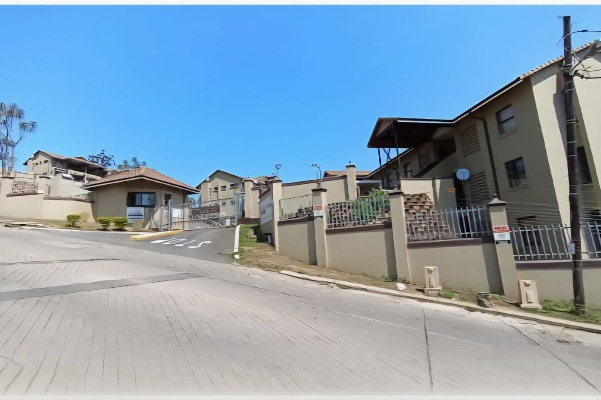 Street view of the townhouse complex entrance featuring a paved driveway, perimeter walling with electric fencing, and a security gatehouse.