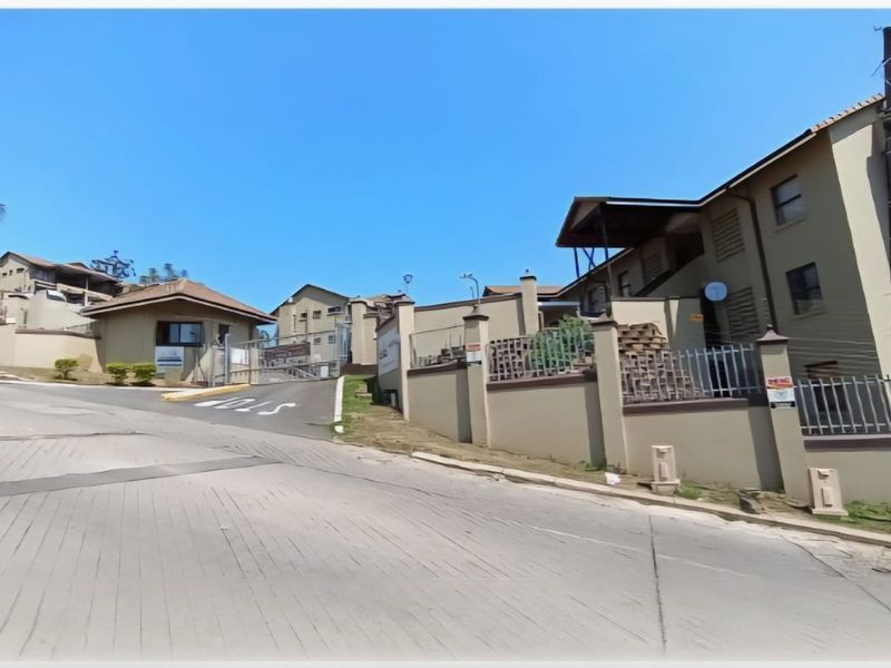 Street view of the townhouse complex entrance featuring a paved driveway, perimeter walling with electric fencing, and a security gatehouse.