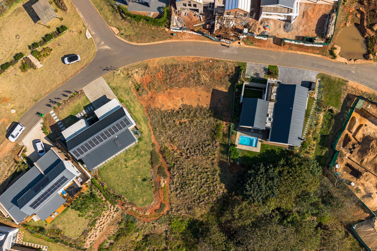 Aerial view of a vacant residential plot situated between established homes in a developed estate, featuring road access and surrounding greenery.