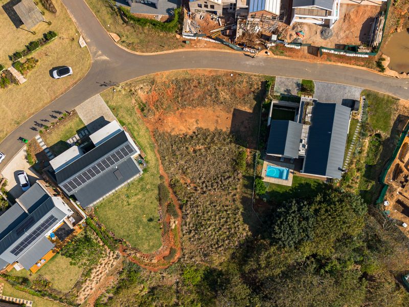 Aerial view of a vacant residential plot situated between established homes in a developed estate, featuring road access and surrounding greenery.