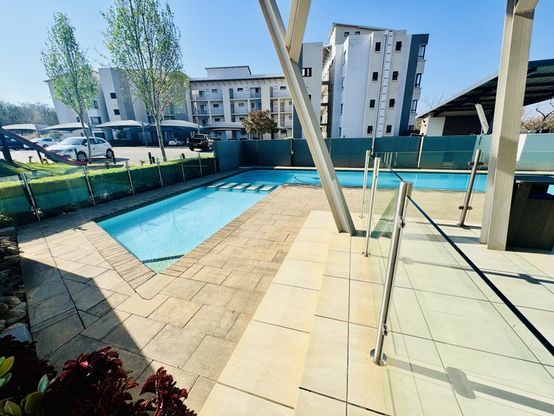 A well-maintained communal swimming pool area featuring paved decking, glass balustrades, and a covered patio structure within a modern apartment complex.