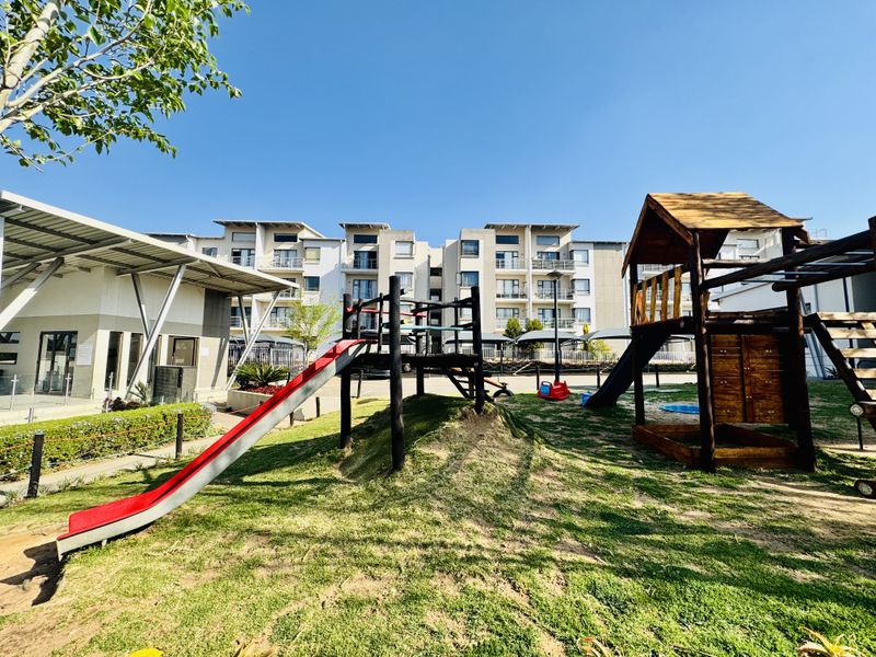 A well-maintained communal garden area featuring a children's playground with slides and climbing structures, set against a modern residential apartment building.