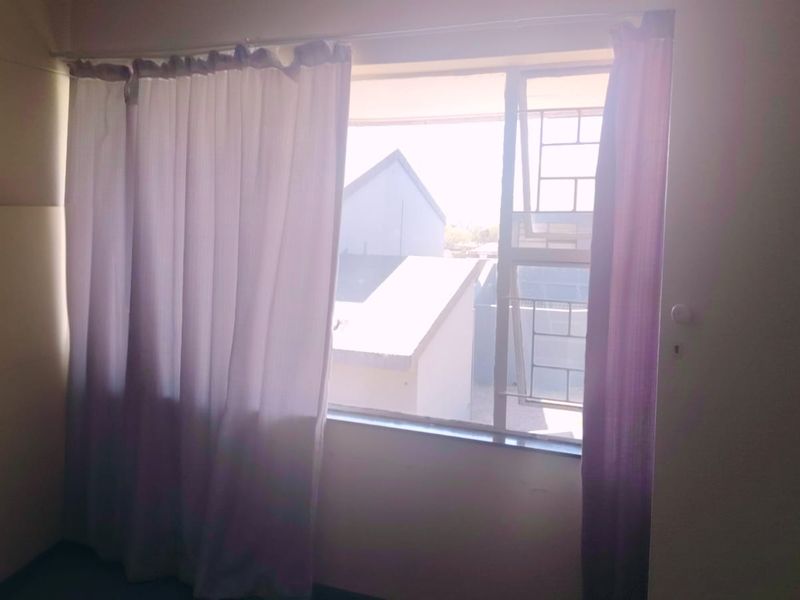 A simple, empty bedroom featuring a window with security bars and a curtain rail, offering a view of the surrounding neighborhood.